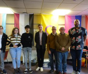 Group of seven people standing in a row with LGBT+ flags behind.