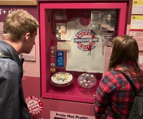 Image of two people looking into a museum cabinet containing a t shirt, badges and other objects.