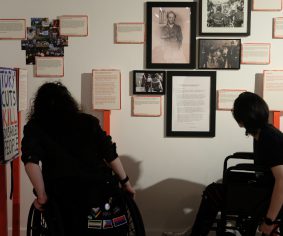 Two people, both using wheelchairs, look at images in an exhibition.